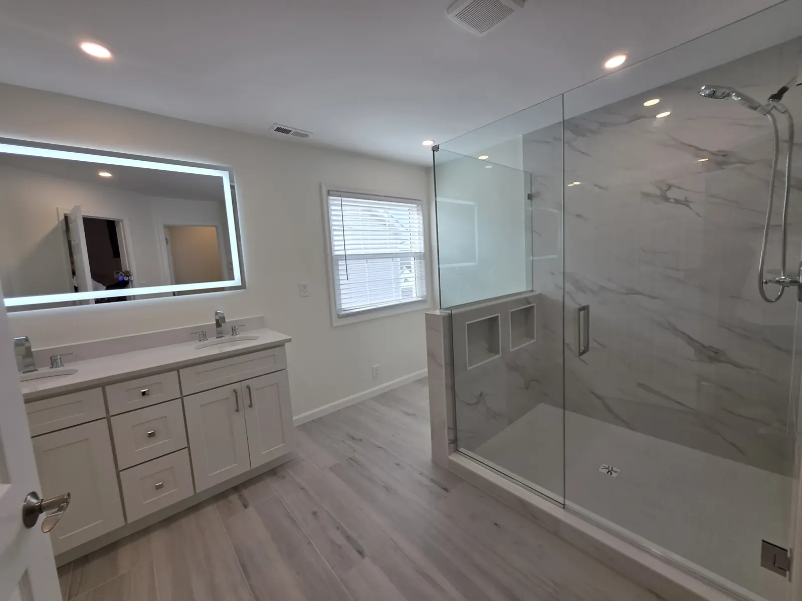 White shaker double vanity with a lit mirror beside a glass shower